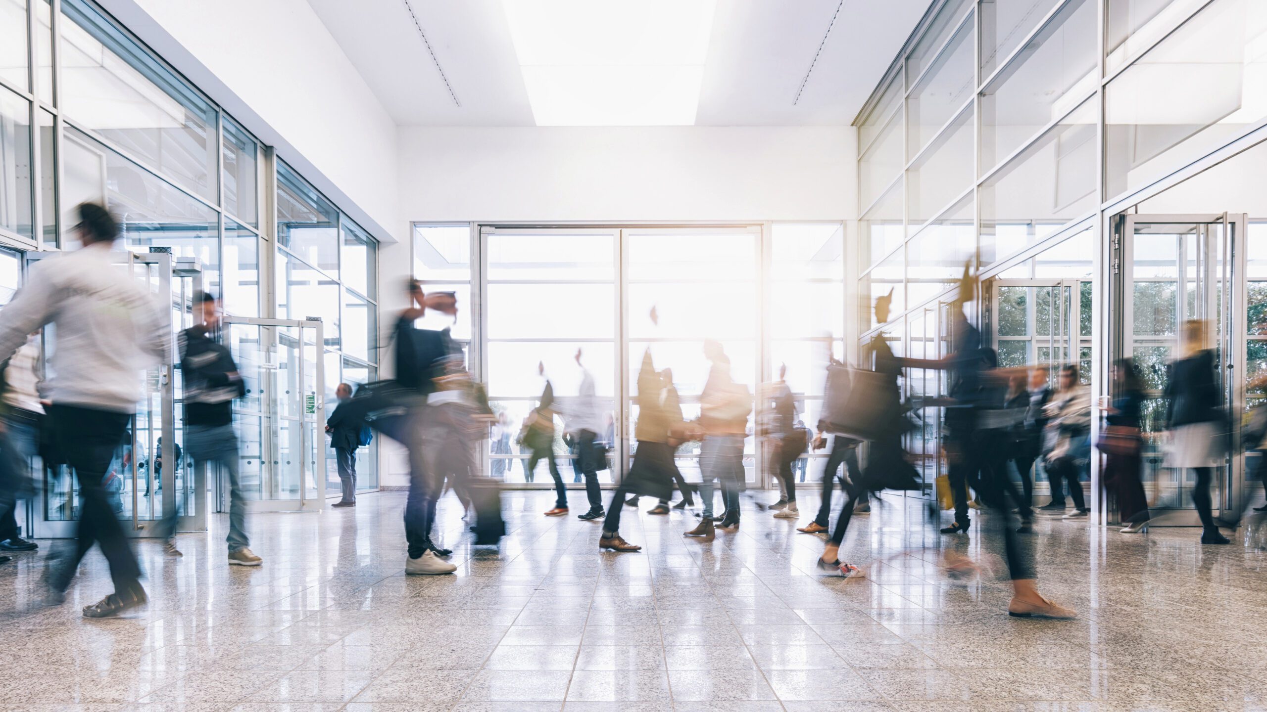 Bewegungsszene in einem modernen, hellen Gebäude mit Glaswänden: Viele Personen gehen in unterschiedliche Richtungen durch eine große Eingangshalle. Die Menschen sind teilweise unscharf dargestellt, was Dynamik und Alltagstrubel vermittelt. Die Szene steht für Mobilität, Arbeitsalltag und gesellschaftliche Bewegung.