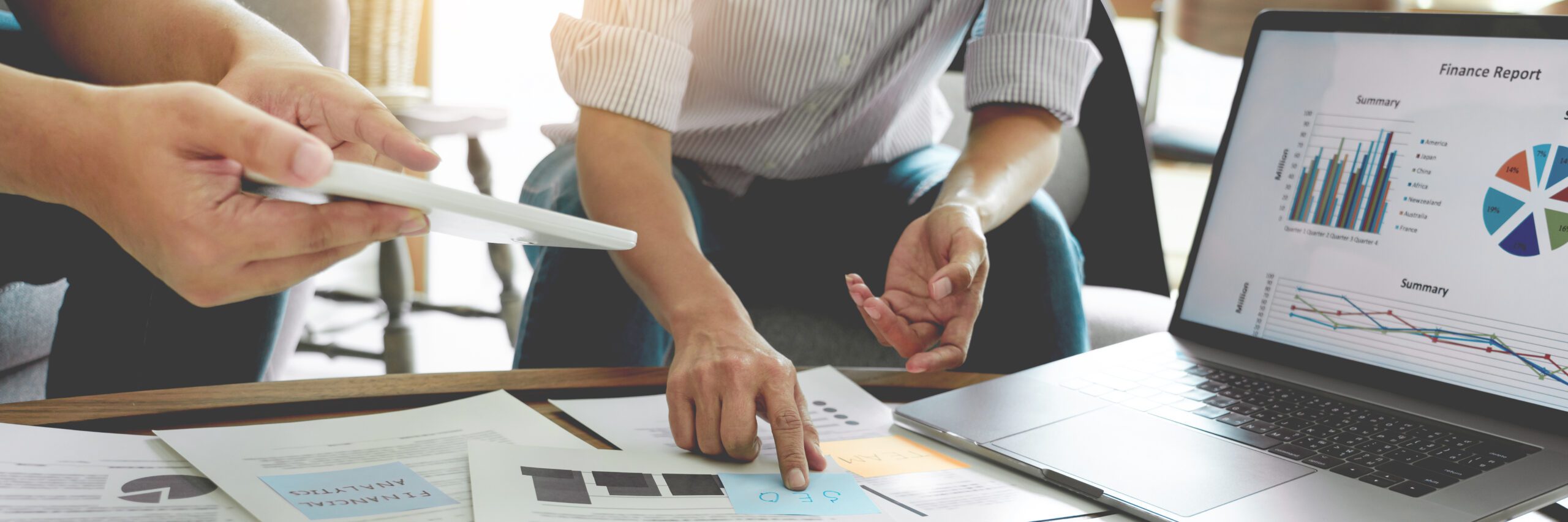 Detailaufnahme einer Arbeitssituation: Zwei Personen sitzen an einem Tisch und besprechen Unterlagen mit Diagrammen und Kennzahlen. Eine Hand zeigt auf ein Dokument, während daneben ein Laptop mit einem Finanzbericht und verschiedenen Diagrammen geöffnet ist. Die Szene steht für Analyse, Planung und gemeinsame Entscheidungsfindung.