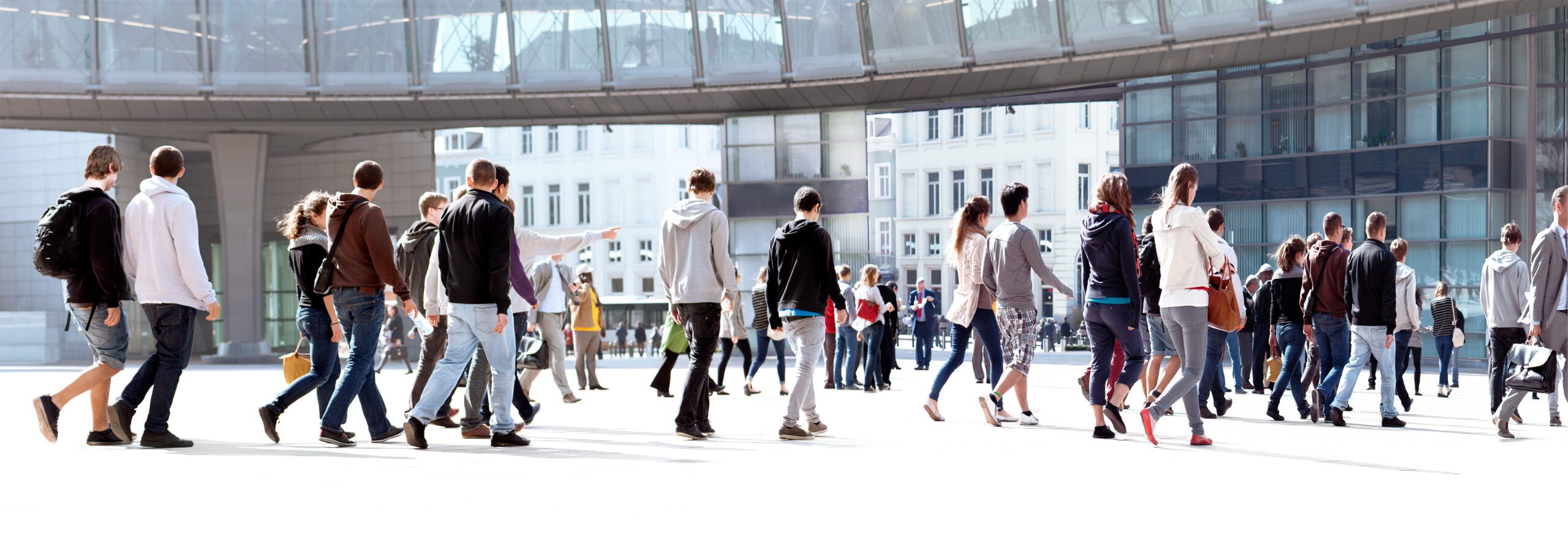 Weitwinkelaufnahme einer größeren Menschengruppe, die über einen offenen, modernen Platz mit Glas- und Bürogebäuden im Hintergrund geht. Die Personen bewegen sich in unterschiedliche Richtungen, tragen Alltagskleidung und Rucksäcke. Die Szene vermittelt Urbanität, Bewegung und öffentliches Leben im Stadtraum.