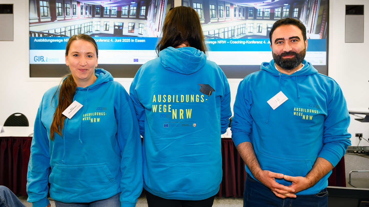 Das Bild zeigt drei junge Menschen, die einen blauen Pulli mit der Aufschrift Ausbildungswege NRW tragen. Die Person in der Mitte ist nur von hinten zu sehen.