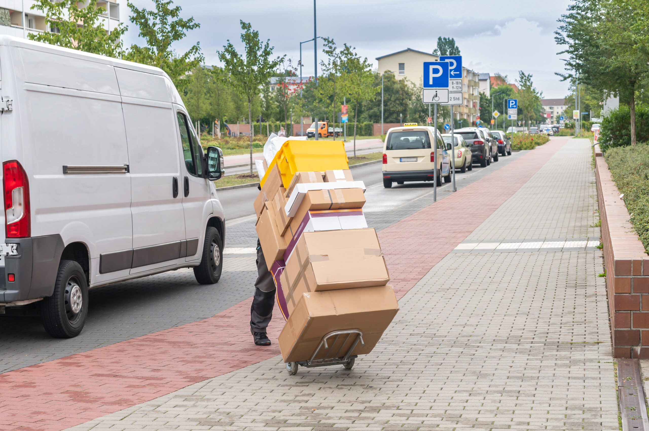 Vor einem Kleintransporter schiebt auf dem Gehweg ein Zusteller einen Rollwagen mit zahlreichen Paketen