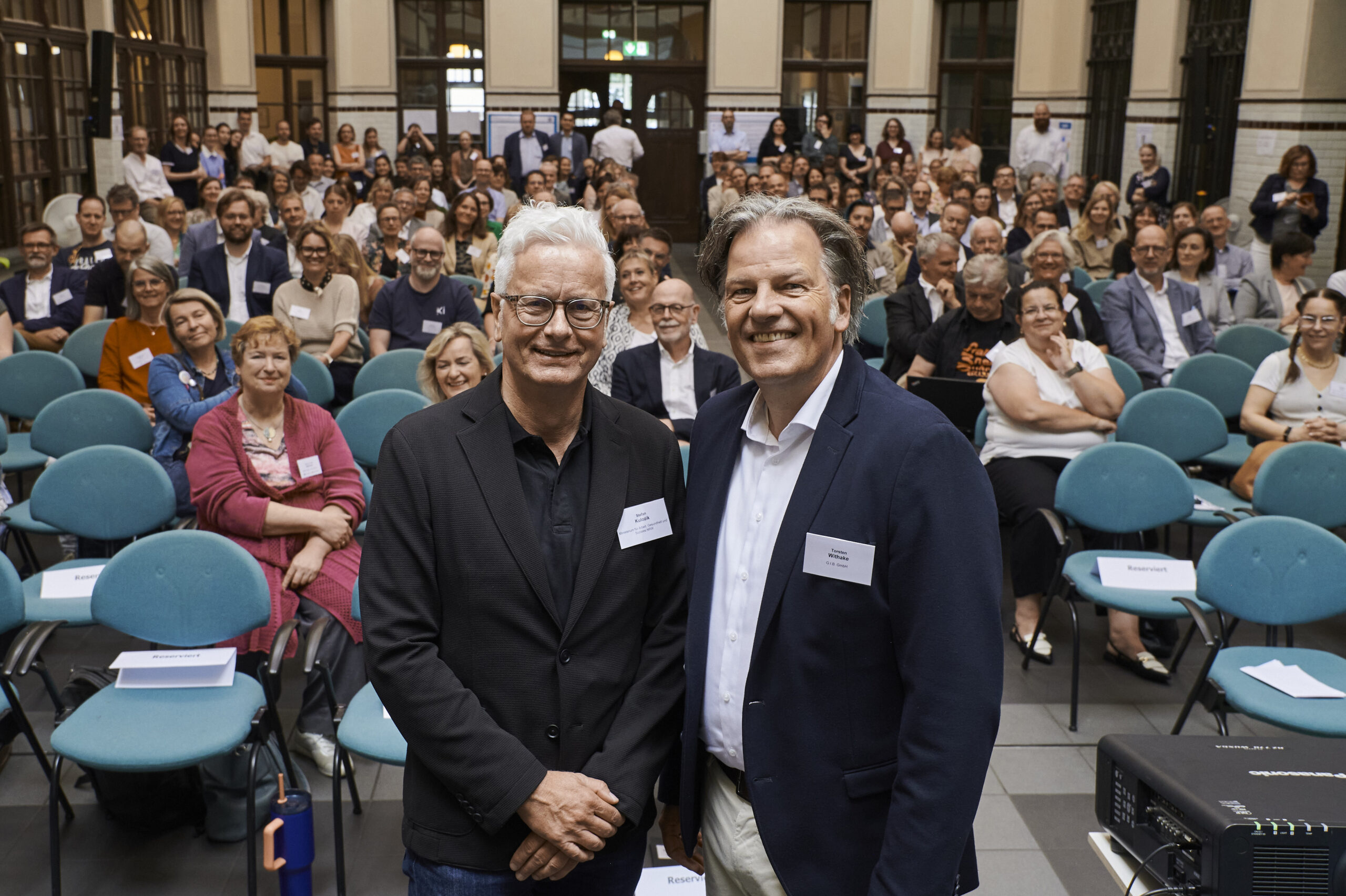 von links: Leiter der Abteilung Arbeit des MAGS NRW neben Geschäftsführer der G.I.B. Torsten Withake stehen in der Veranstaltungshalle vor dem Publikum.